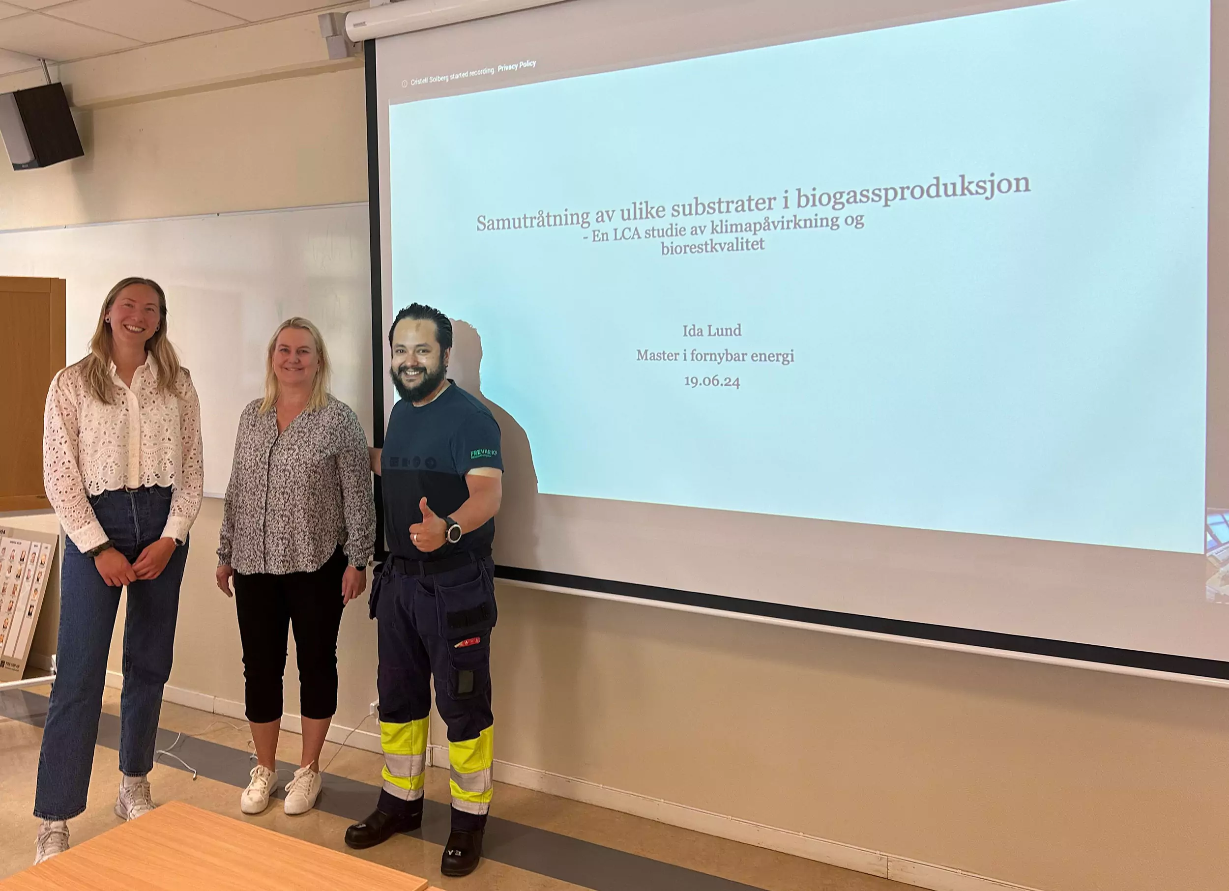 To kvinner og en mann, som viser tommel opp, står ved siden av et lærret med en presentasjon av en studentoppgave. Masteroppgave skrevet om fornybar energi ved FREVAR.