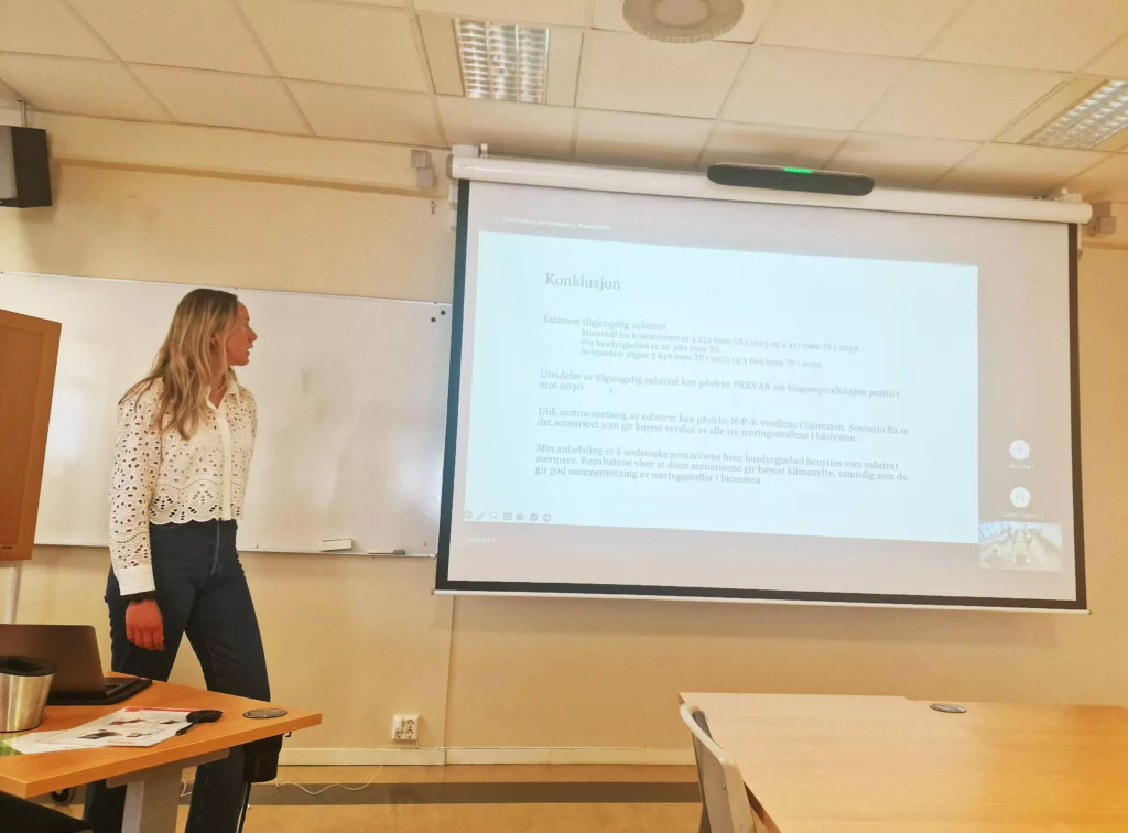 Digital presentasjon på som vises et lærret i et møterom. En ung kvinne står til venstre for presentasjonen og snakker med ansiktet vendt mot sin presentasjon.