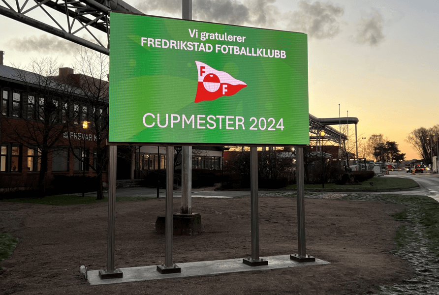 Infoskilt utenfor et bygg. Skiltet har grønn bakgrunn med teksten: Vi gratulerer FREDRIKSTAD FOTBALLKLUBB - CUPMESTER 2024.