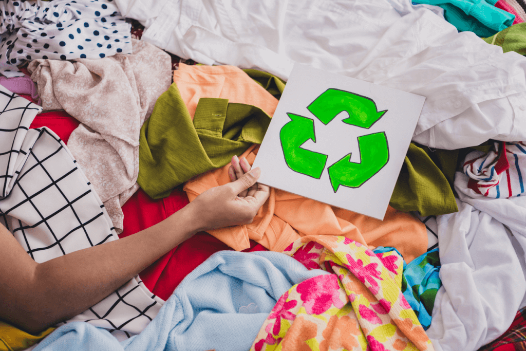 En hånd som holder et hvitt arke med en grønt resirkulerings ikon over en haug av forskjellige tekstiler og klær.
