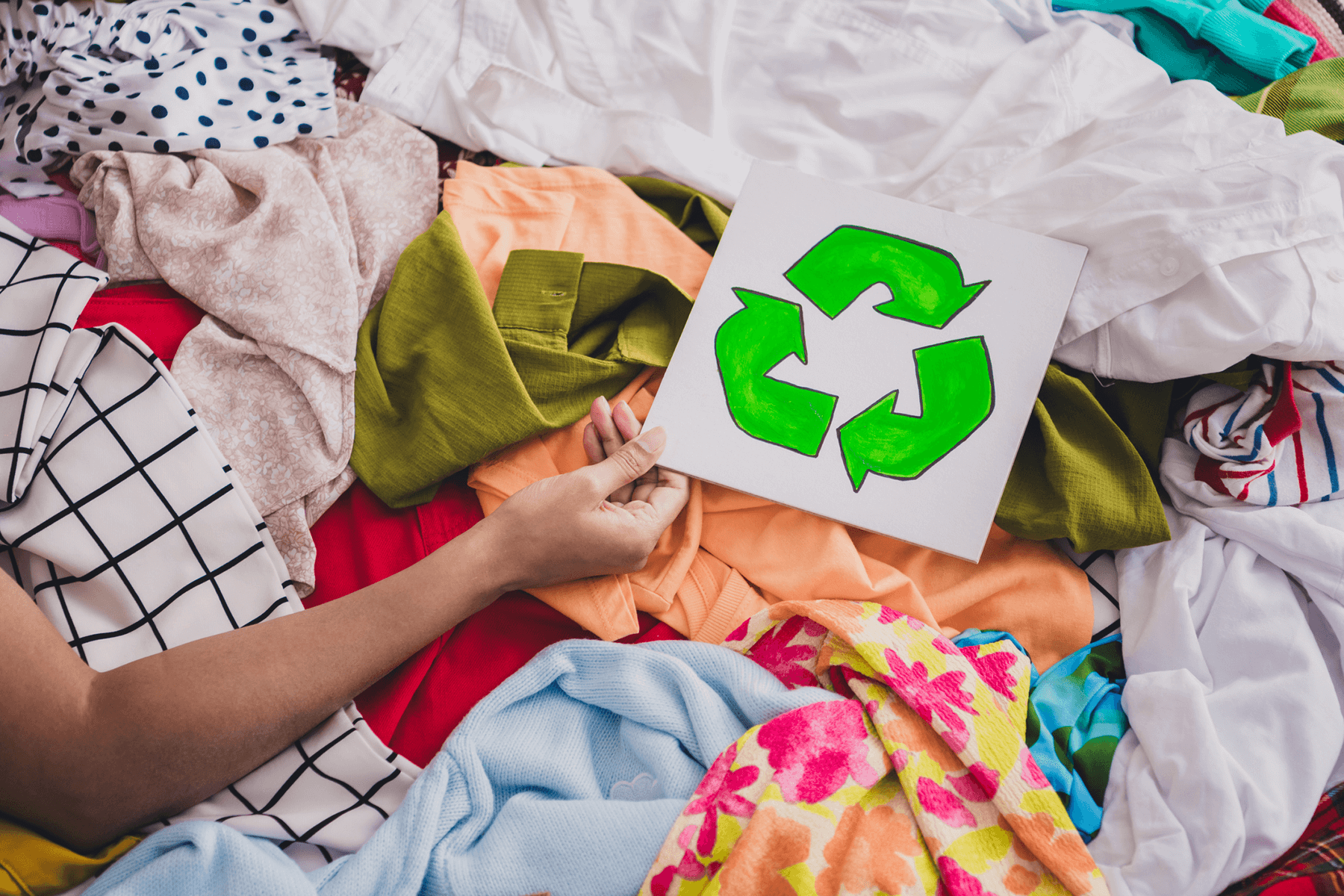 En hånd som holder et hvitt arke med en grønt resirkulerings ikon over en haug av forskjellige tekstiler og klær.