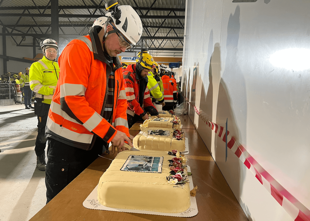 Arbeidere med verneutstyr som hjelm og jakke, står i kø for å dele opp og spise fire marsipankaker dekorerte med blomster og bilde av et bygg. 