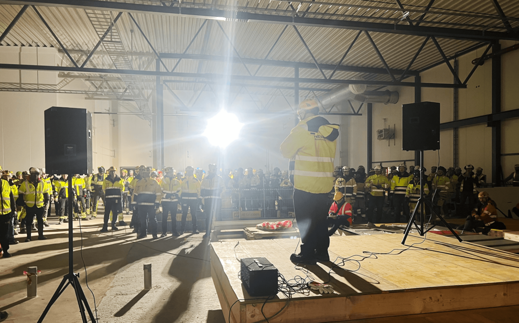 Samling av arbeidere i et åpent areal i et industribygg. En mann står på et podium i forkant og snakker ut mot forsamlingen. Alle har på seg verneutstyr som hjelmer og jakker.