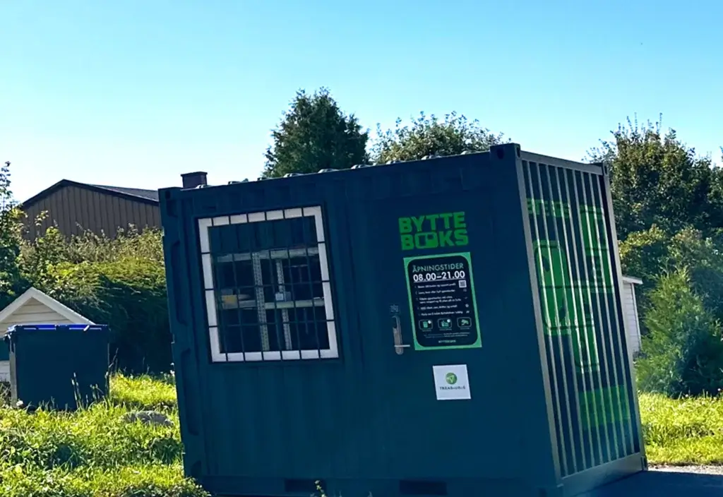 Et kontainerhus i blått med "bytteboks" skrevet på. Kontaineren har et vindu med gitter og står utendørs omgitt av gress, busker og trær under blå himmel.