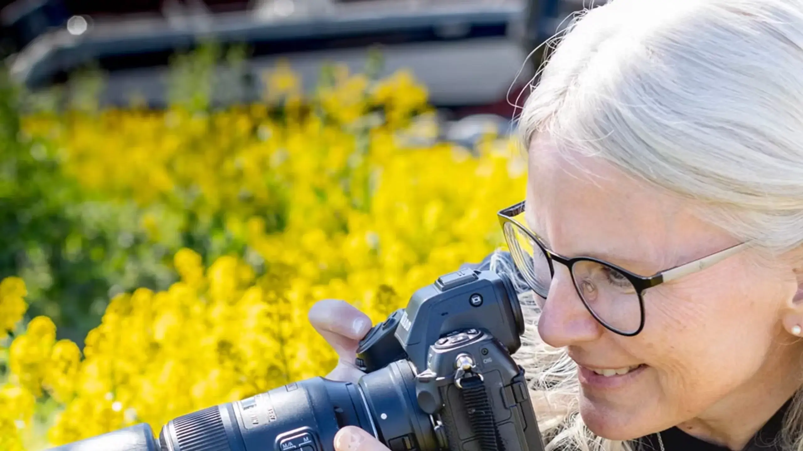n kvinne med lyst hår og briller smiler mens hun ser gjennom søkeren på et kamera. I bakgrunnen er det en uskarp eng med gule blomster.